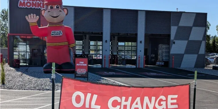 A Grease Monkey auto service center celebrates its Grand Opening, featuring a large inflatable mascot and a red banner reading "OIL CHANGE" in front of the building.