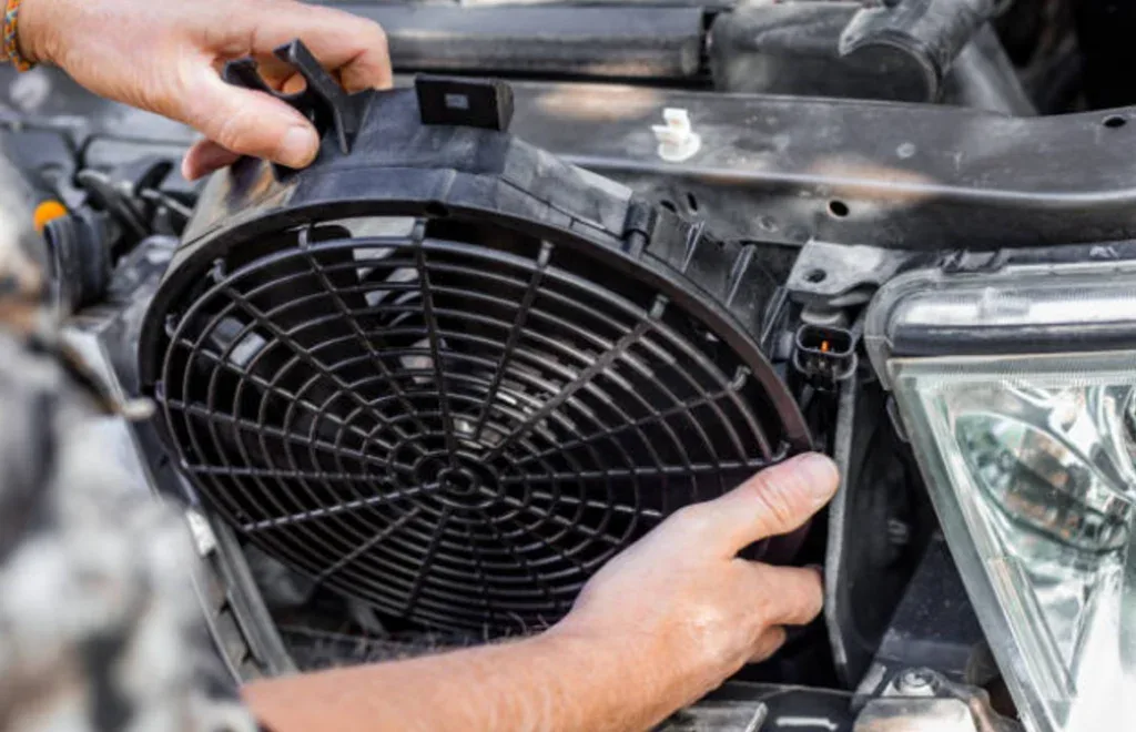 A person installs or removes a car radiator fan near the engine bay and headlight area as part of vehicle air conditioning or AC system service in Aurora, Colorado.