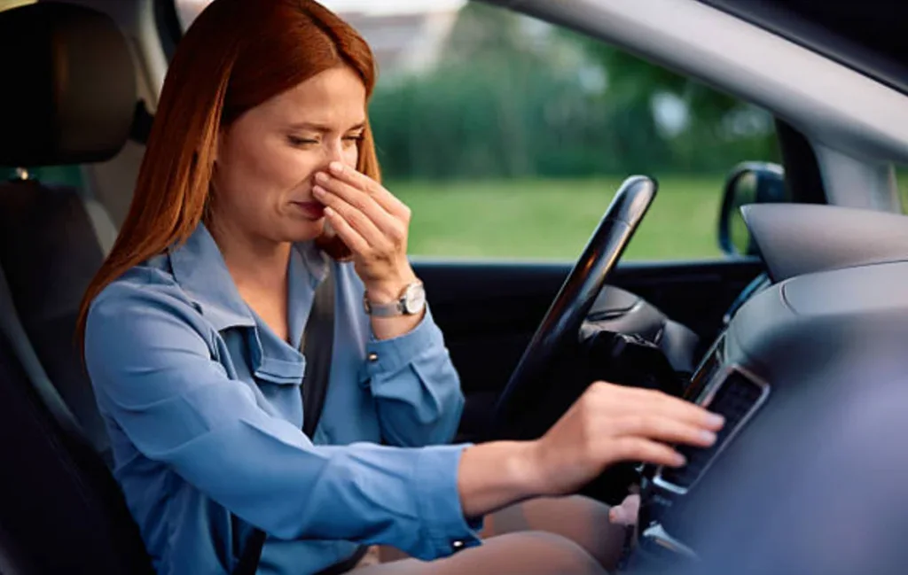 A woman sitting in the driver's seat of a car in Laramie, Wyoming pinches her nose and adjusts the vehicle’s air conditioning, appearing bothered by an unpleasant smell.