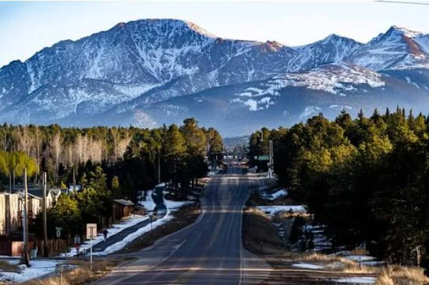 A paved road winds through a forested area near Woodland Park Colorado, leading toward tall, snow-capped mountains under a clear sky—perfect for enjoying a drive with your vehicle air conditioning or getting reliable AC service nearby.