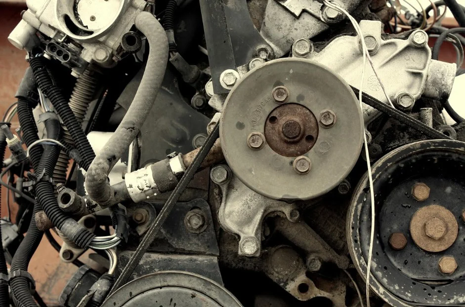 Close-up of a used car engine in Southeast Pocatello, showing metal pulleys, belts, wires, and various mechanical components with visible dirt and wear—reminding drivers about the importance of regular vehicle air conditioning service.