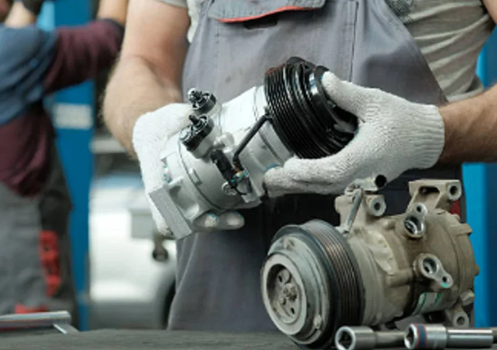A mechanic in gloves holds a new car air conditioning compressor, with an old unit and tools on the workbench—showcasing expert Vehicle Air Conditioning Service in the heart of Kimberly, Idaho, serving the greater Magic Valley area.