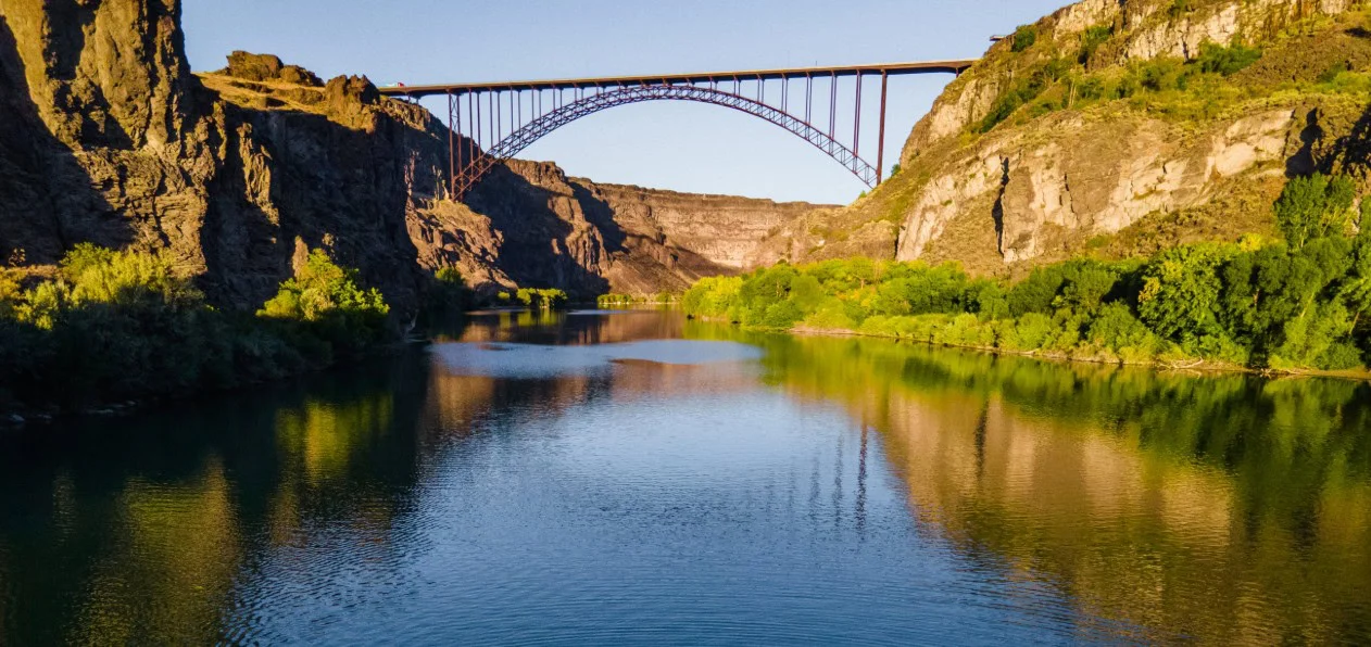 A steel arch bridge spans a deep canyon over a calm river, with rocky cliffs and green vegetation on both sides under a clear sky.