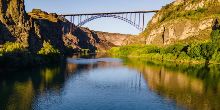 A steel arch bridge spans a deep canyon over a calm river, with rocky cliffs and green vegetation on both sides under a clear sky.