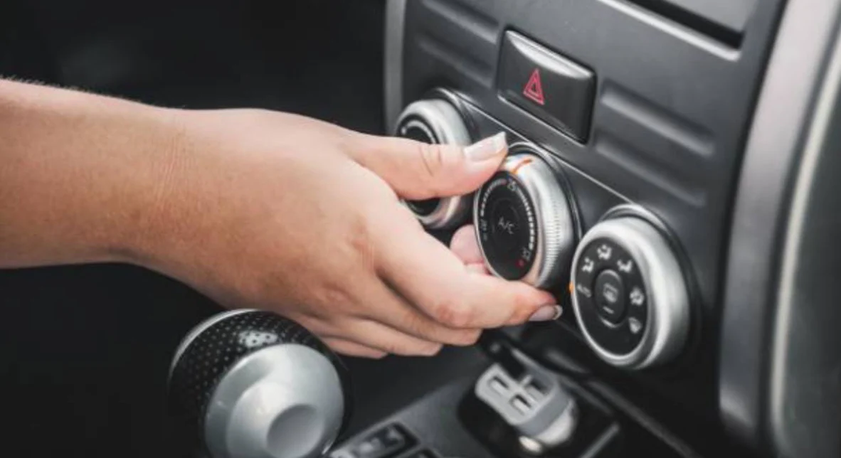 A person adjusts the air conditioning control knob on a car dashboard, with the gear shift and hazard light button visible nearby.