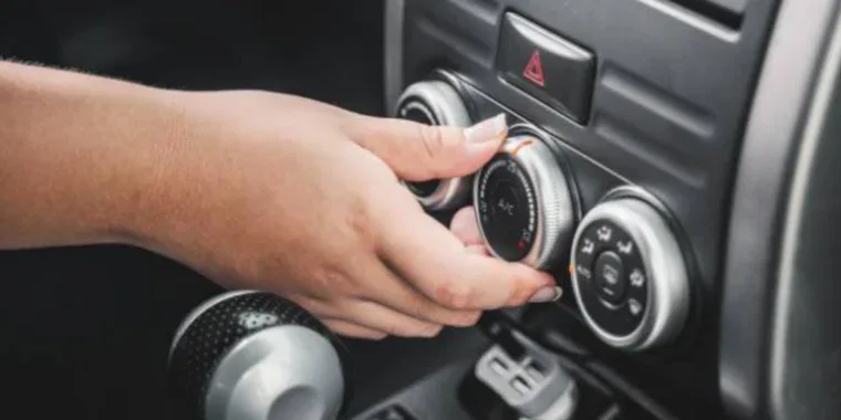 A person adjusts the air conditioning control knob on a car dashboard, with the gear shift and hazard light button visible nearby.