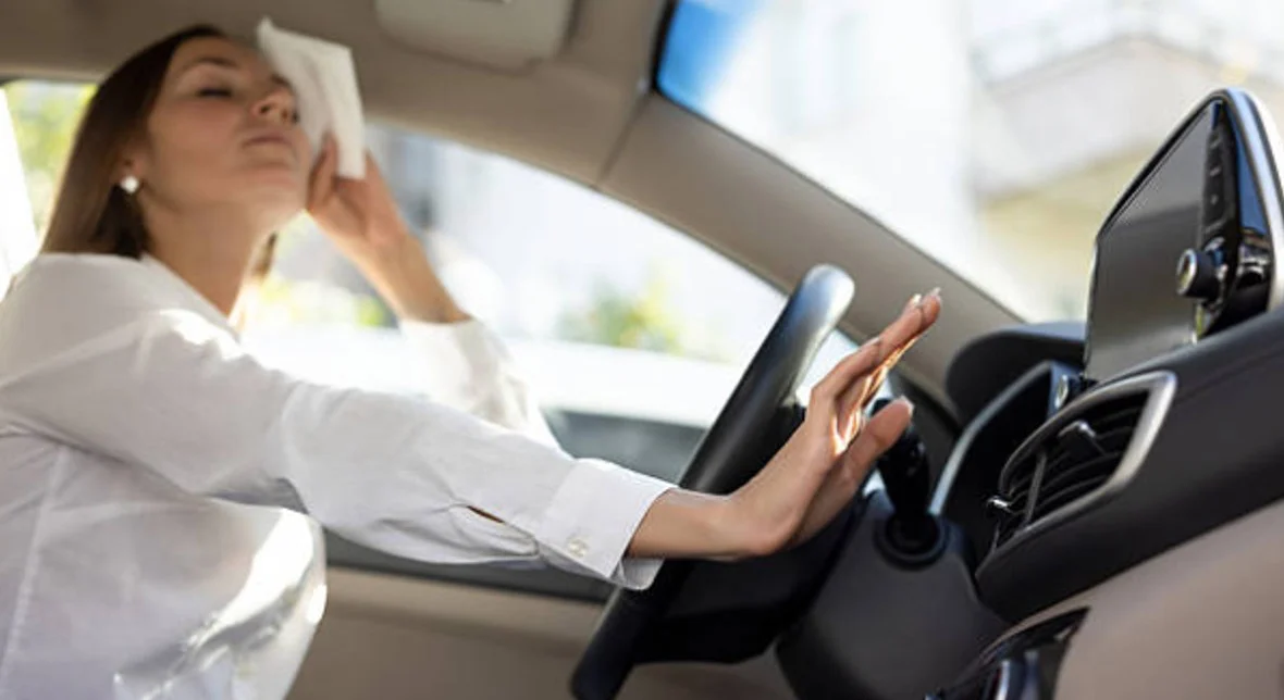 A woman sits in a car, wiping her forehead with a tissue and reaching to adjust the air conditioning controls on the dashboard.