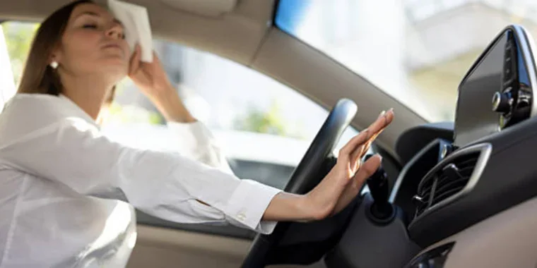 A woman sits in a car, wiping her forehead with a tissue and reaching to adjust the air conditioning controls on the dashboard.
