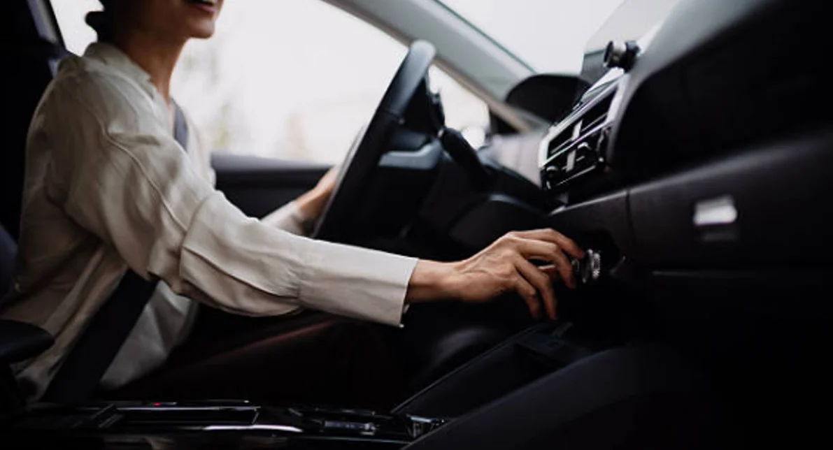 Person sitting in a car driver's seat, adjusting a control knob on the dashboard, with one hand on the steering wheel.