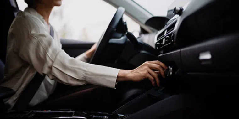Person sitting in a car driver's seat, adjusting a control knob on the dashboard, with one hand on the steering wheel.
