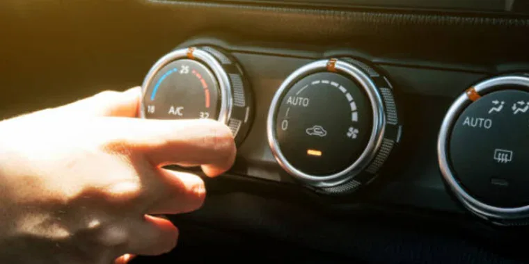 A hand adjusts the temperature dial on a car's air conditioning control panel, with two other dials for airflow and fan speed visible.
