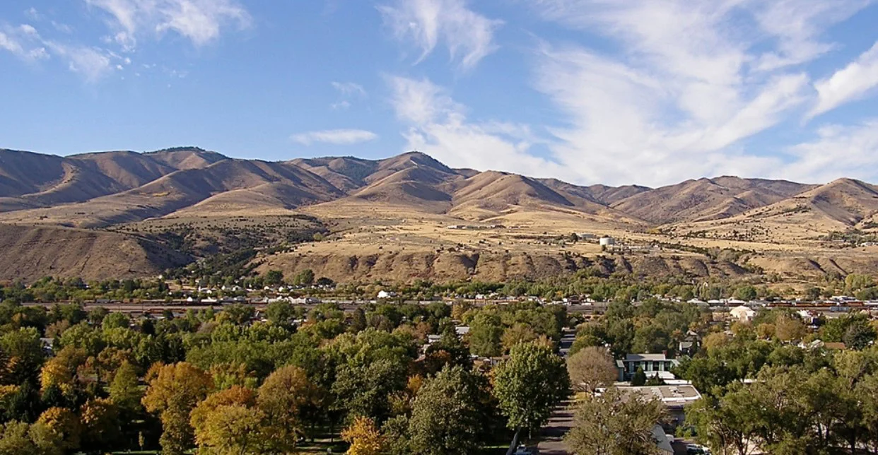 A view of Pocatello, Idaho, with green trees in the foreground and dry, brown hills under a blue sky with wispy clouds in the background—a perfect day to think about vehicle air conditioning and summer car maintenance.