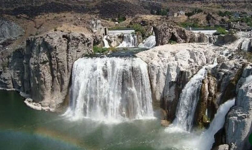 Large waterfall cascading over rocky cliffs into a river in Twin Falls, Idaho, with a faint rainbow at the base and buildings visible in the background.