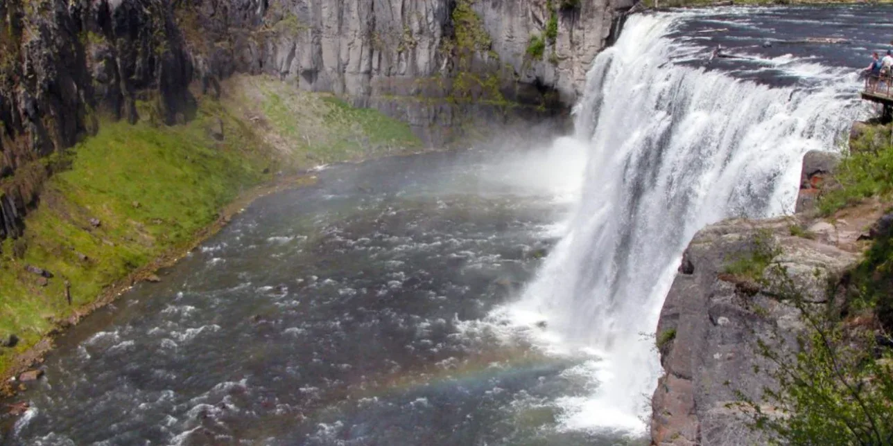 Wide waterfall cascading over a rocky ledge into a river below near Rexburg, Idaho, with mist rising and a faint rainbow at the water’s surface. Lush greenery surrounds this serene scene.