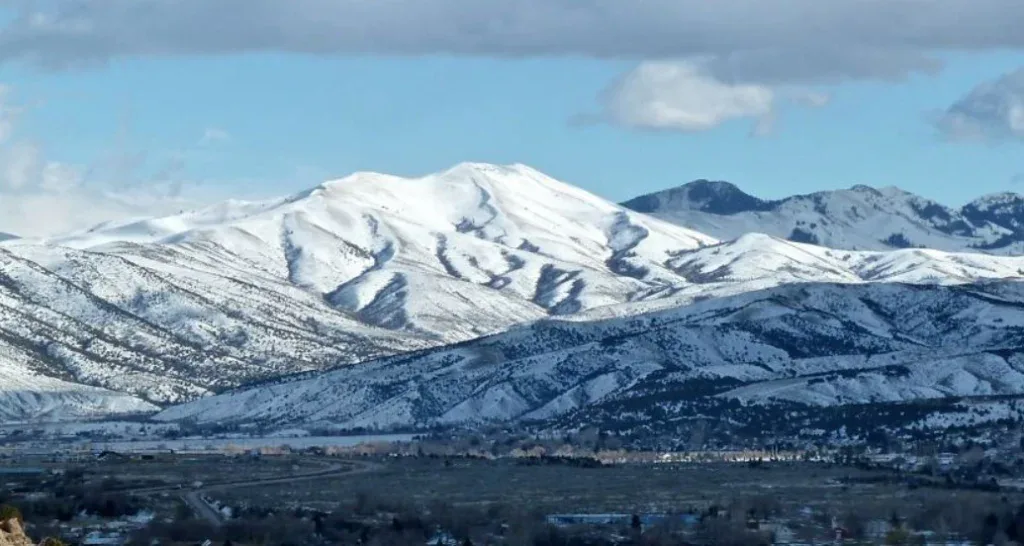Snow-covered mountains with a partly cloudy sky overlook Southeast Pocatello, where drivers navigate lower hills and a valley dotted with scattered trees, buildings, and trusted vehicle air conditioning service centers at the base.