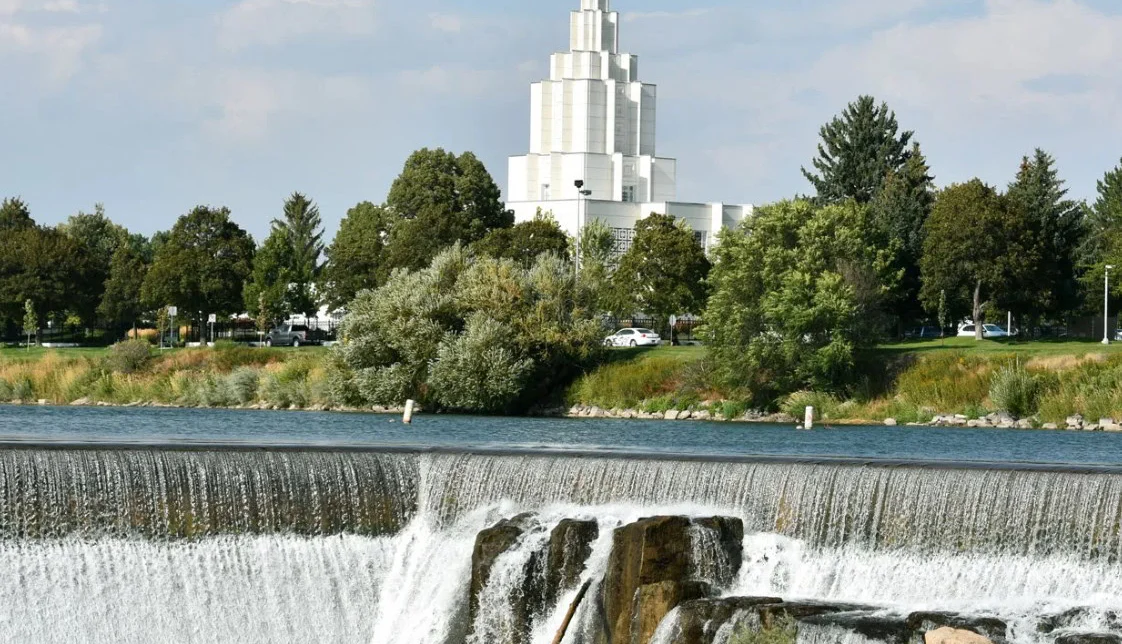 A white temple in Idaho Falls with a gold statue atop its spire stands behind a row of trees and a cascading river waterfall under a partly cloudy sky.