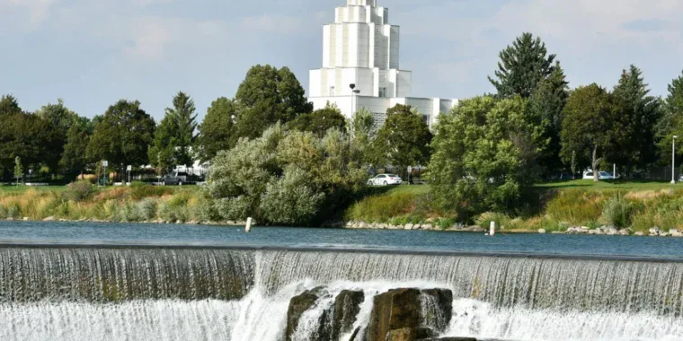 A white temple in Idaho Falls with a gold statue atop its spire stands behind a row of trees and a cascading river waterfall under a partly cloudy sky.