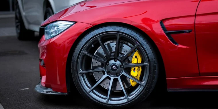Close-up of the front left wheel of a red sports car with a high-performance brake system featuring yellow brake calipers, parked on a dark paved surface next to a silver vehicle in Aurora, Colorado.
