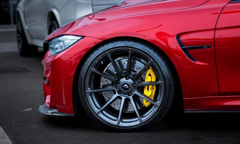 Close-up of the front left wheel of a red sports car with a high-performance brake system featuring yellow brake calipers, parked on a dark paved surface next to a silver vehicle in Aurora, Colorado.
