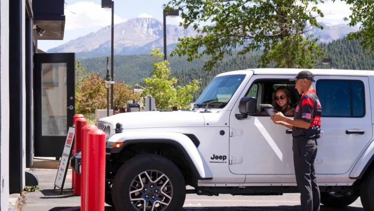 A person in a uniform assists a woman in a white Jeep at a drive-thru with mountains and trees visible in the background.