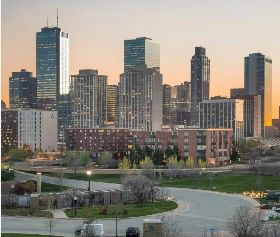 City skyline with modern high-rise buildings at sunset, a curving road in the foreground, and some trees and grass visible—an ideal setting to remind drivers of the importance of regular vehicle maintenance, including timely brake service in Aurora, to ensure safe journeys through urban landscapes.