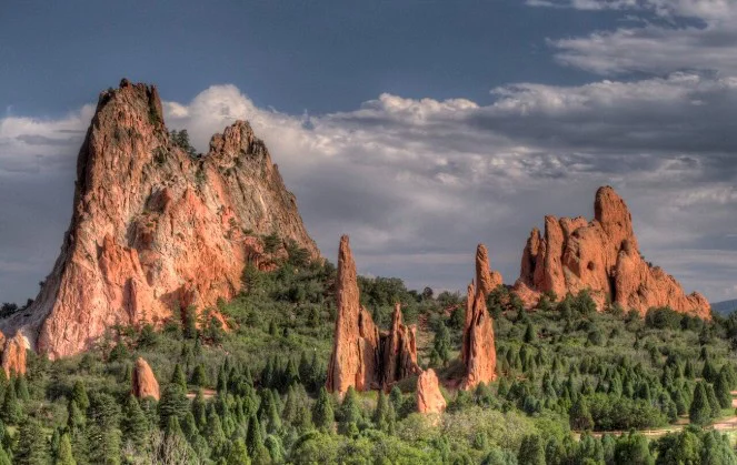 Jagged red rock formations rise above green trees under a partly cloudy sky at Garden of the Gods park, one of Colorado Springs’ most stunning natural landmarks.