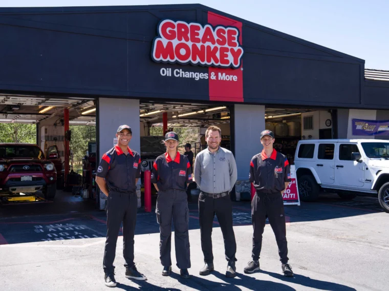 Four employees stand in front of a Grease Monkey auto service center, one of many convenient Front Range locations, with two vehicles parked in open service bays behind them on a sunny day.