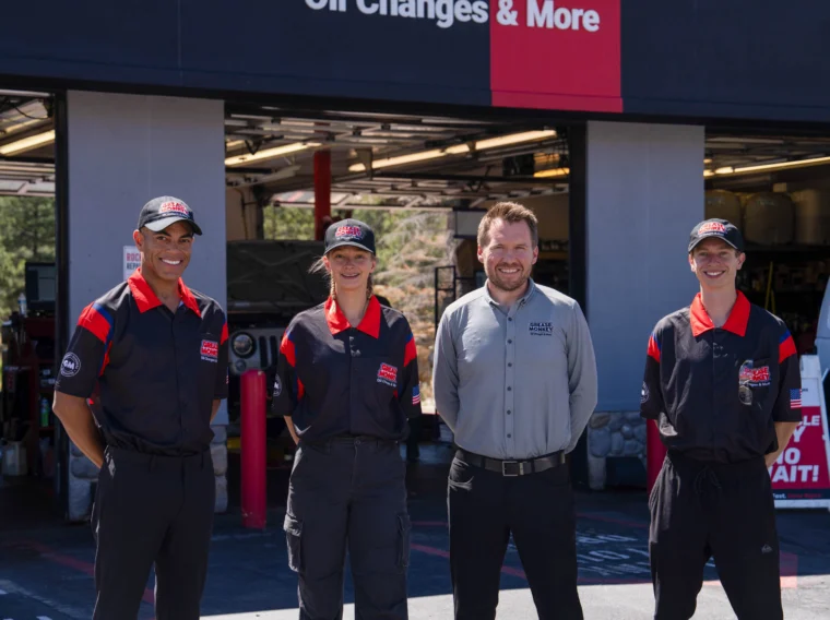 Four employees stand in front of an auto service garage, three in black and red uniforms and one in a gray shirt, smiling at the camera.
