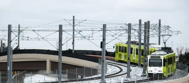A light rail train with green cars travels on snowy tracks beside a fenced overpass under cloudy skies in Aurora, Colorado, where regular vehicle maintenance and brake system service are essential for safe winter journeys.