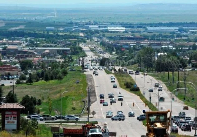 A wide view of a busy road in Colorado Springs with cars, businesses, and some construction vehicles in the foreground, surrounded by greenery and distant buildings.