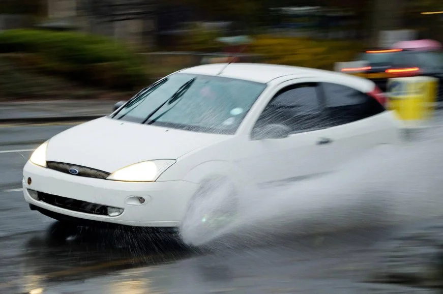 A white car drives quickly through a large puddle on a wet road in Aurora, Colorado, splashing water to the side—a vivid reminder of the importance of maintaining your brake system and seeking regular brake service in Aurora.
