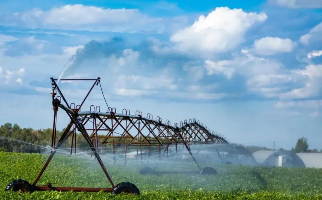A large irrigation system sprays water over a green crop field under a partly cloudy sky, with farm buildings—including those reminiscent of the agricultural landscape near Rexburg Idaho—visible in the background.