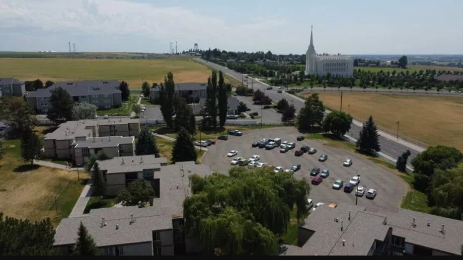 Aerial view of an apartment complex with parked cars, open fields, and the Yellowstone Highway Corridor running alongside a main road. In the background, a white temple-like building stands under a partly cloudy sky near Rexburg, with power transmission lines visible in the landscape.