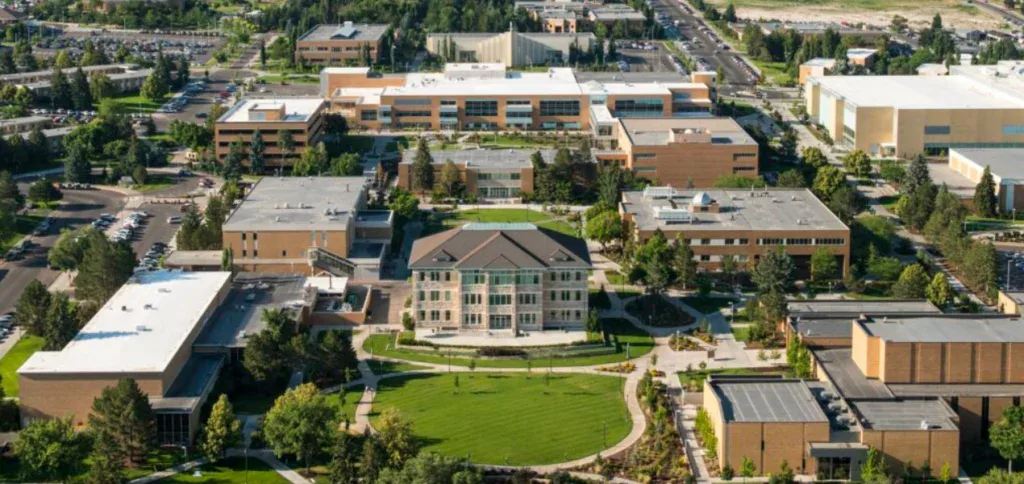Aerial view of the Rexburg university campus with multiple academic buildings, green lawns, walking paths, trees, and parking lots stretching alongside the Yellowstone Highway Corridor under a clear sky.