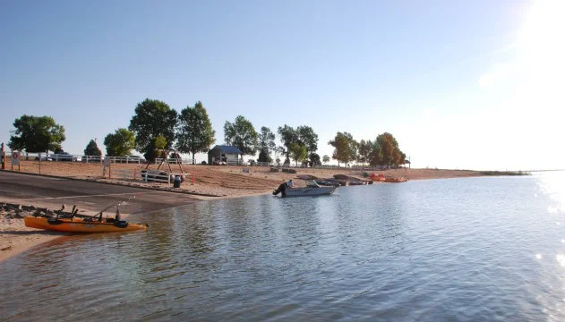 A lakeshore in Aurora, Colorado, with a boat ramp, parked vehicles, boats in the water, kayaks on the sand, and trees under a clear blue sky—easily accessible from I-225 and perfect for relaxing while staying alert for any transmission emergency updates.