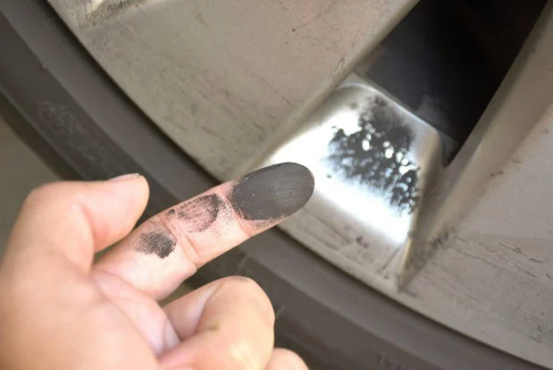 A hand points to black brake dust on a car wheel, with a finger covered in the same dust—a clear sign that your brake system may need attention. If you’re in Woodland Park, consider scheduling a vehicle service to keep your brakes performing safely.