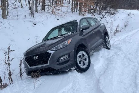 A gray SUV, likely facing transmission problems, is stuck in a snowy ditch beside a road lined with bare trees.