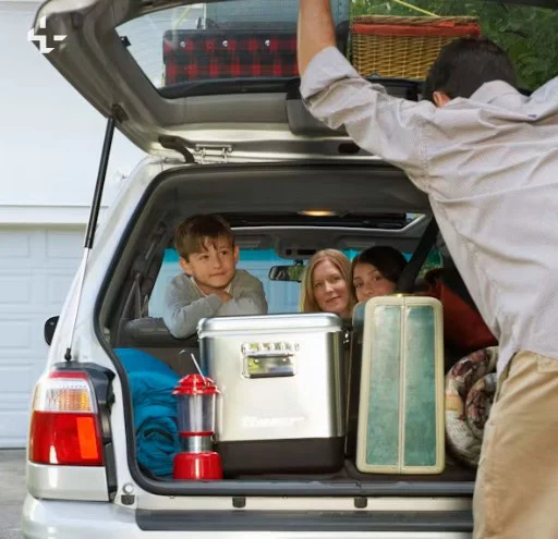 A family sits in the back of an open SUV packed with luggage, a cooler, a lantern, and camping supplies, preparing for a trip through Woodland Park along scenic Ute Pass—carefully checking for any transmission warning signs before their adventure begins.
