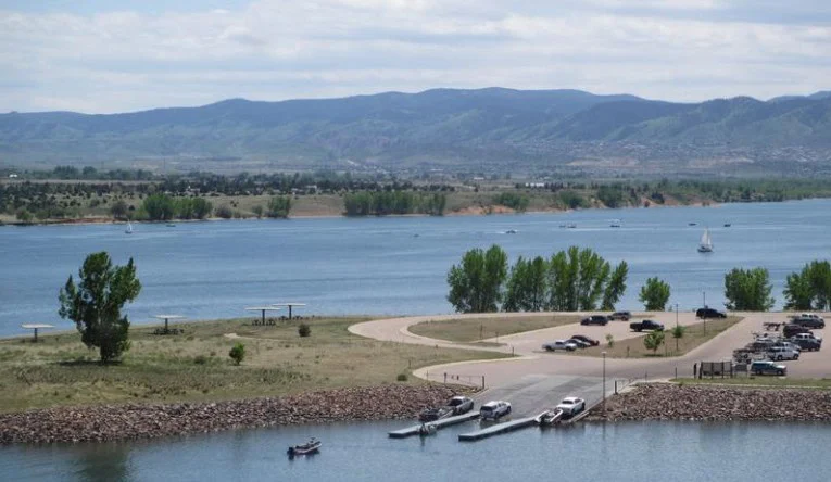 A lake in Littleton, Colorado with several boats, a boat ramp, parked cars, and distant mountains under a partly cloudy sky.