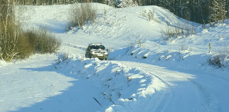 A car drives along a snow-covered, winding road through a rural landscape with snow-covered trees and hills in the background—a scene familiar to many Littleton drivers who know to watch for transmission warning signs and be mindful of potential transmission problems, especially in challenging winter conditions.