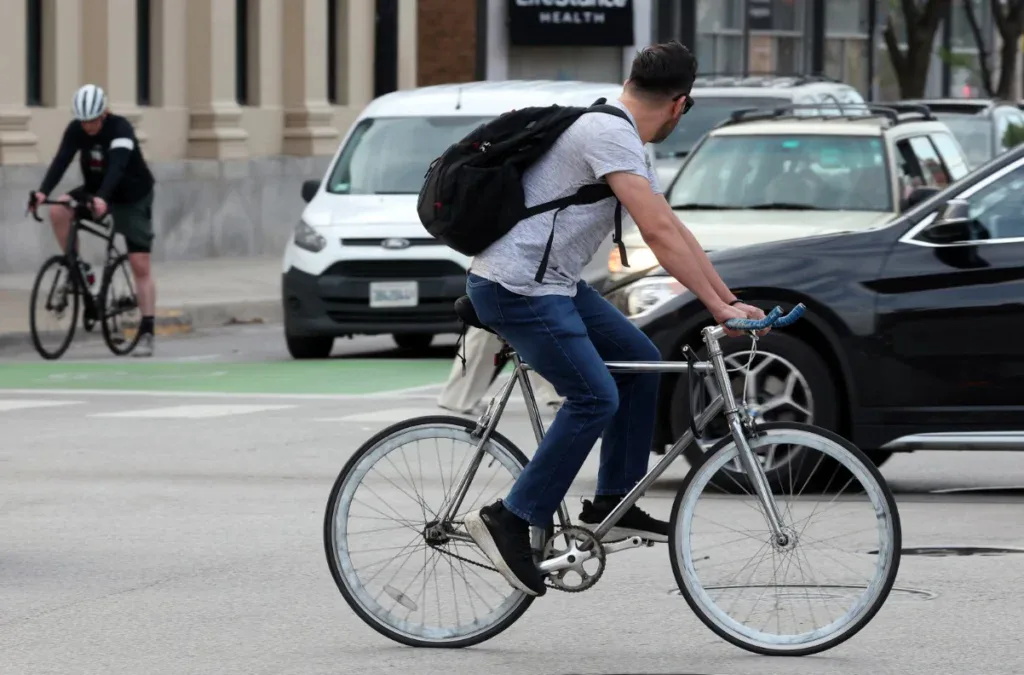 A person wearing jeans and a backpack rides a bicycle through a city street; another cyclist and several cars—perhaps some heading to Grease Monkey Pocatello for vehicle maintenance—are visible in the background.