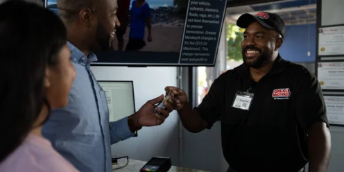 A smiling employee hands car keys to a customer at a service counter while another person stands nearby. A television and certificates are visible in the background.