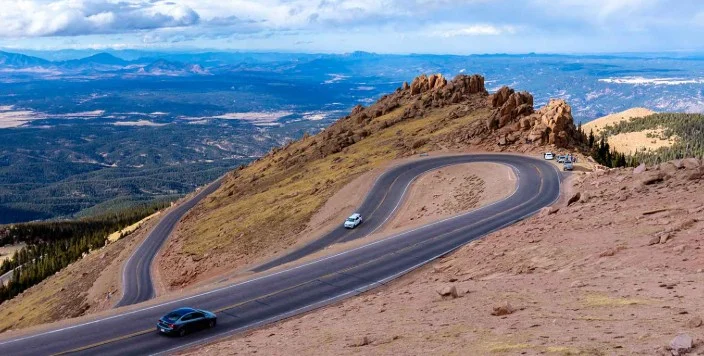 Curving mountain road with several cars driving near Colorado Springs, rocky terrain in foreground, and distant forested landscape under a partly cloudy sky.