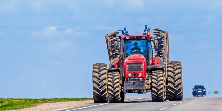 A red tractor with large dual tires drives on a rural road near Kimberly, Idaho, with a car approaching in the opposite lane under a blue sky.