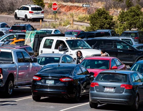 Crowded parking lot with various cars and trucks parked closely together near Fort Carson. Two people walk between vehicles, and a stop sign is visible in the background.