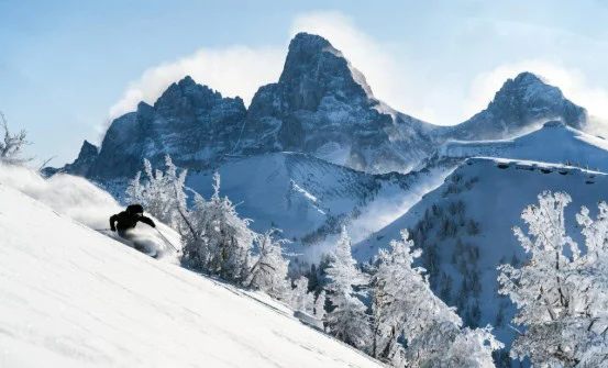 A skier moves downhill through fresh snow along the Yellowstone Highway Corridor, with jagged, snow-covered mountain peaks and frosted trees in the background under a clear sky.