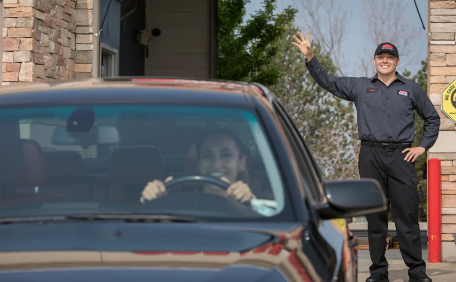 A person smiles and waves while standing outside a building in Pocatello as a car with a driver approaches, both appearing happy and engaged—perhaps pleased after receiving regular service for the vehicle’s brake system.