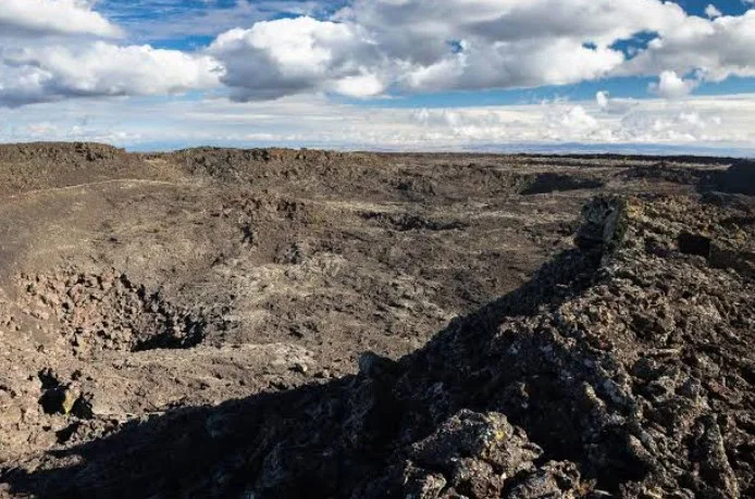 Rocky volcanic landscape under a partly cloudy sky, featuring uneven dark lava rocks and sparse vegetation near Kelly Canyon, North Idaho Falls.