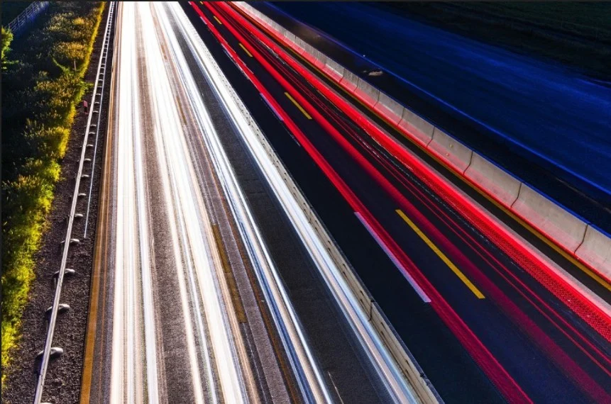 Long-exposure photo of a highway at night showing white and red light trails from passing vehicles, with a guardrail and road markings visible—a reminder of the importance of vehicle maintenance and expert brake system service offered by Grease Monkey Rexburg.