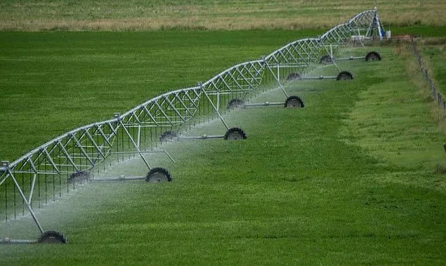 A center-pivot irrigation system sprays water over a large, green agricultural field near Idaho Falls, not far from where transmission lines stretch across the landscape to support the daily INL commute.